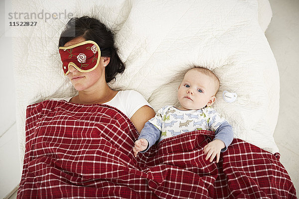Mutter mit Schlafmaske und wachem Baby im Bett liegend