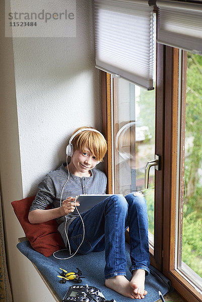 Junge sitzt auf der Fensterbank mit digitalem Tablett und trägt Kopfhörer.