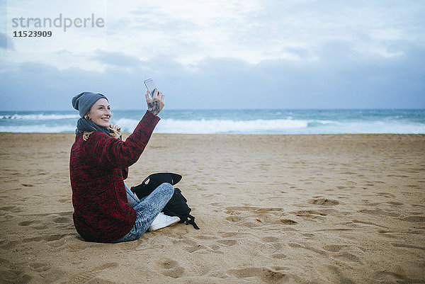 Lächelnde junge Frau  die im Winter am Strand sitzt und ihr Handy benutzt.