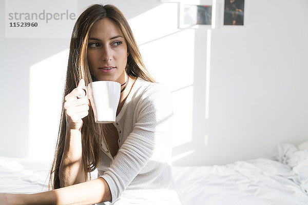 Junge Frau sitzt auf dem Bett und hält eine Tasse Kaffee.