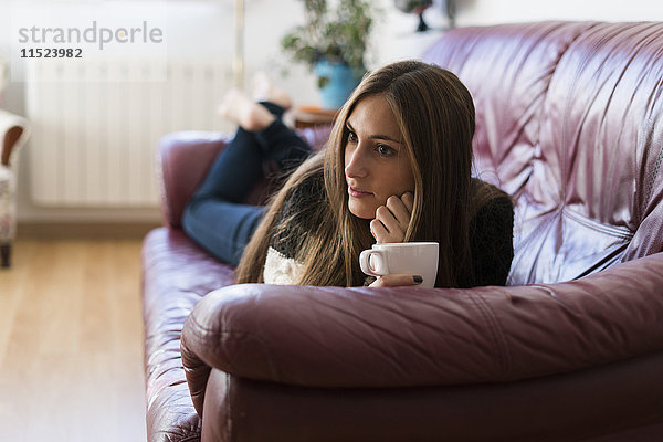 Junge Frau liegt auf der Couch und hält eine Tasse Kaffee.
