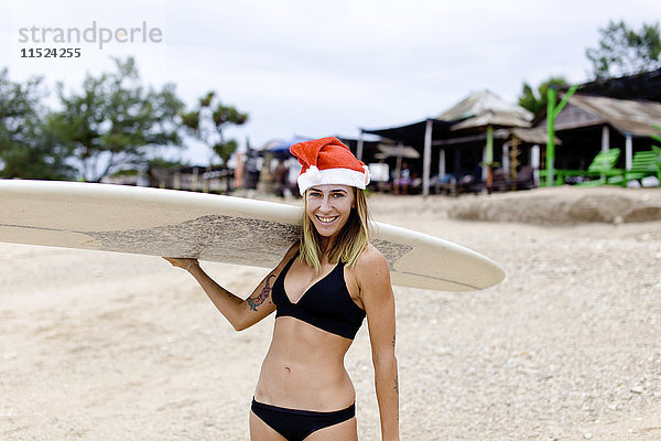 Indonesien  Bali  lächelnde Frau mit Surfbrett am Strand mit Weihnachtsmütze