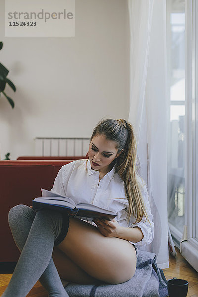 Junge Frau sitzt auf dem Boden des Wohnzimmers und liest ein Buch.