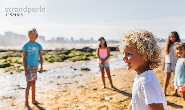 Fröhlicher kleiner Junge am Strand mit anderen Kindern im Hintergrund
