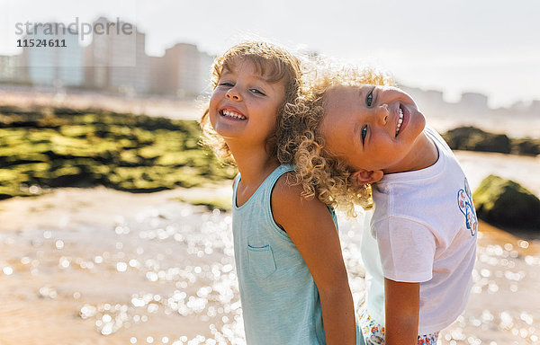 Porträt eines glücklichen kleinen Jungen und Mädchens  das Rücken an Rücken am Strand steht.