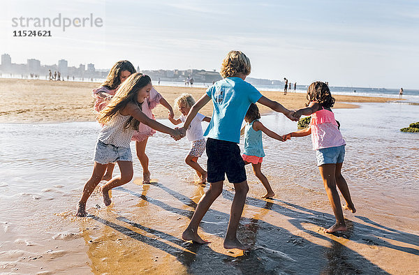 Gruppe von sechs Kindern  die am Strand Ring-a-ring-a-roses spielen.