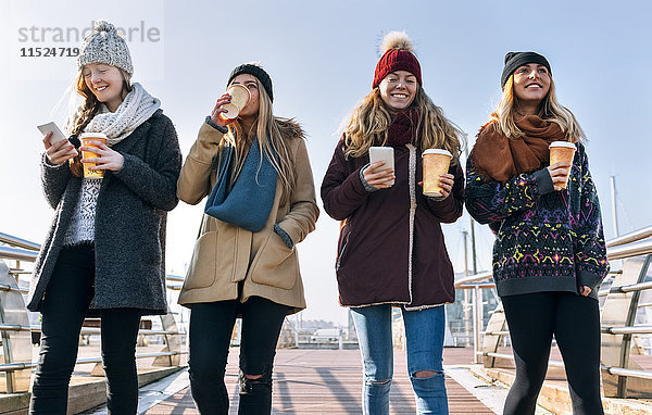 Vier Freunde mit Kaffee zum Mitnehmen und Handys  die im Winter auf einer Brücke spazieren gehen.