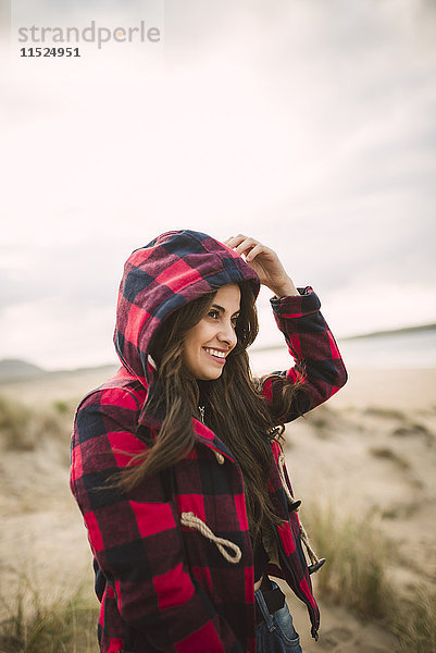 Lächelnde junge Frau mit langen braunen Haaren in Kapuzenjacke am Strand