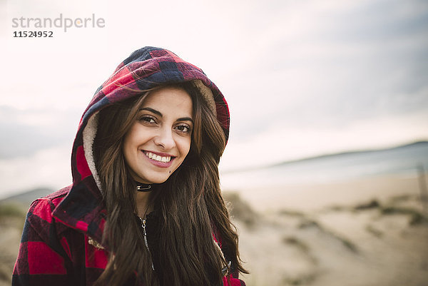 Porträt einer lächelnden jungen Frau in Kapuzenjacke am Strand