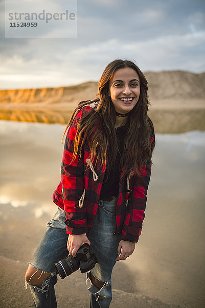 Porträt einer lächelnden jungen Frau mit Kamera am Strand