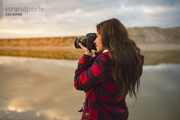 Junge Frau beim Fotografieren am Strand mit Kamera bei Sonnenuntergang