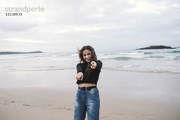 Porträt einer glücklichen jungen Frau am Strand  die auf den Betrachter zeigt.