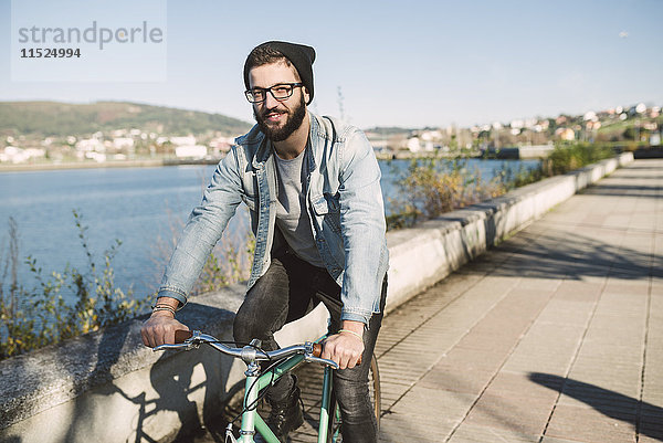 Lächelnder junger Mann auf seinem Fixie-Bike am Wasser