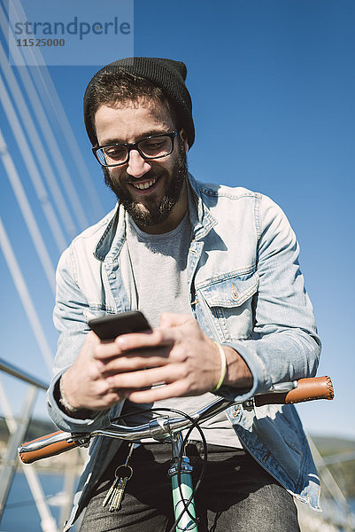 Lächelnder junger Mann mit Fixie Bike mit einem Smartphone auf einer Brücke