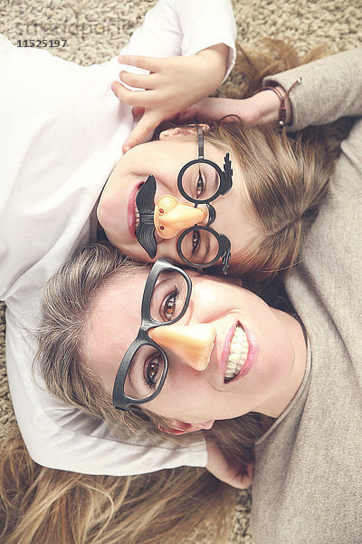 Porträt von Mutter und Tochter auf dem Teppich liegend mit lustiger Brille und Plastiknase