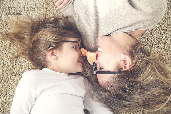Mutter und Tochter liegen auf dem Teppich und tragen eine lustige Brille mit Plastiknase.