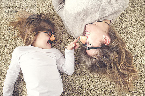 Mutter und kleine Tochter liegen auf dem Teppich und haben Spaß mit Comedy-Brillen