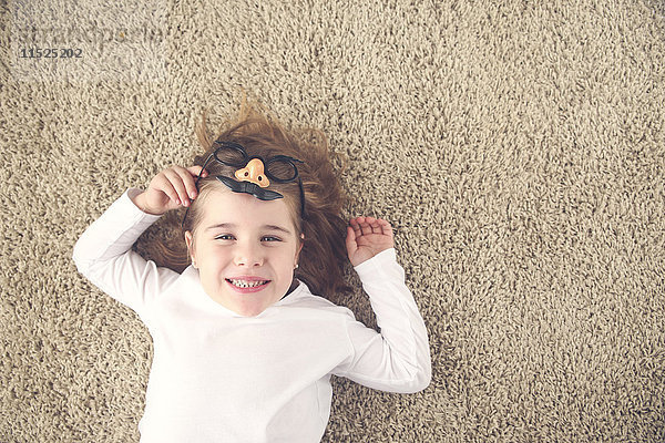 Porträt eines kleinen Mädchens  das auf dem Teppich liegt und eine lustige Brille mit Plastiknase hält.