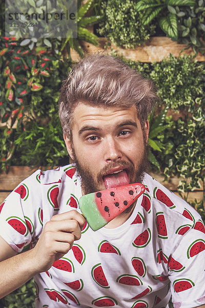 Porträt eines jungen Mannes  der Wassermelonen-Eis-Lolly auf der Terrasse vor dem vertikalen Garten isst.