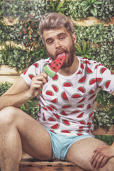 Porträt eines jungen Mannes  der Wassermelonen-Eis-Lolly auf der Terrasse vor dem vertikalen Garten isst.