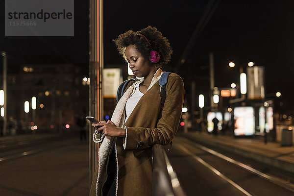 Junge Frau mit Kopfhörer und Smartphone wartet an der Straßenbahnhaltestelle