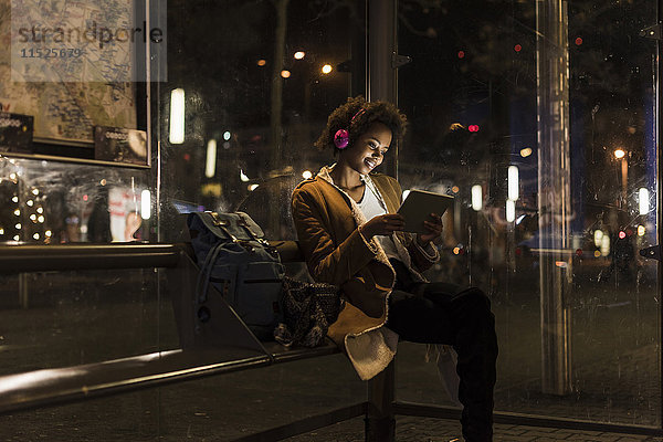 Junge Frau mit Kopfhörer und Tablette wartet an der Straßenbahnhaltestelle