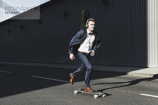 Junger Mann mit Gitarrenkoffer beim Skateboardfahren auf der Straße