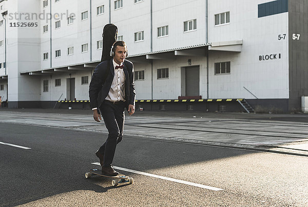 Junger Mann mit Gitarrenkoffer beim Skateboardfahren auf der Straße
