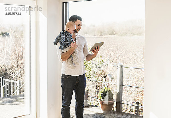 Vater steht zu Hause  trägt Baby-Sohn und liest Textnachrichten