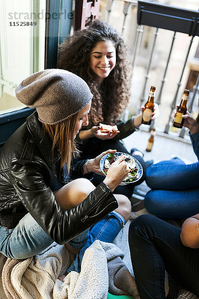 Gruppe von Freunden sitzt auf dem Boden und isst Pizza und Salat und trinkt Bier zu Hause.