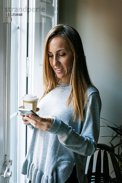 Junge Frau am Fenster mit Handy und Kaffee zum Mitnehmen