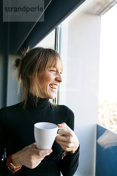 Glückliche junge Frau zu Hause trinkt eine Tasse Kaffee und schaut aus dem Fenster.