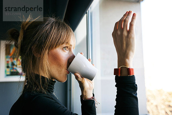 Junge Frau trinkt eine Tasse Kaffee und schaut aus dem Fenster.