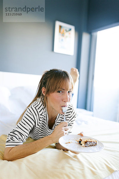 Junge Frau isst ein Stück veganen Kuchen  liegt auf dem Bett  leckt den Löffel.