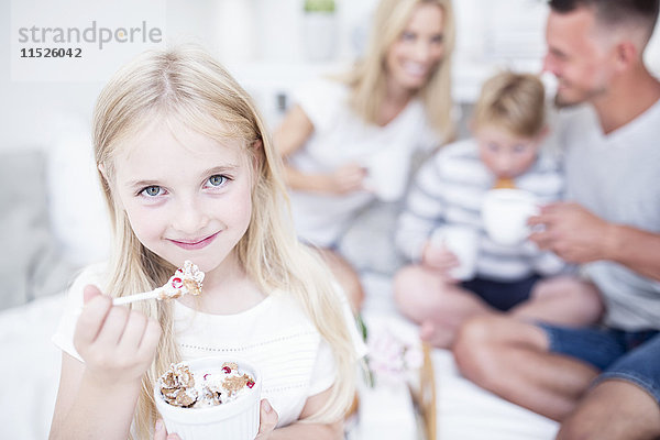 Porträt eines lächelnden Mädchens beim Essen aus der Müslischale mit Familie im Hintergrund
