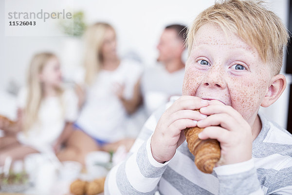 Porträt eines Jungen  der in ein Croissant beißt  mit Familie im Hintergrund