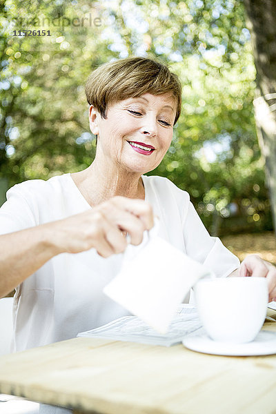 Lächelnde Seniorin gießt Milch in die Tasse Kaffee im Freien