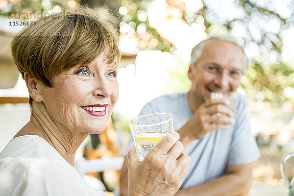 Lächelnde Seniorin trinkt ein Glas Wasser mit Ehemann im Hintergrund