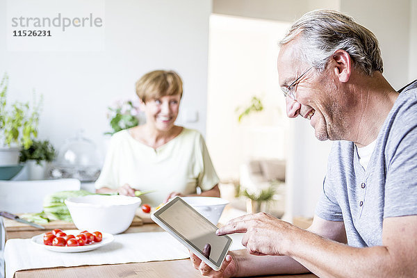 Senior Mann zu Hause mit digitaler Tablette und Frau  die im Hintergrund Salat zubereitet.