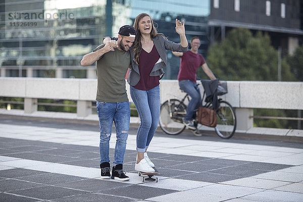 Junger Mann unterstützt lachende Freundin auf Skateboard