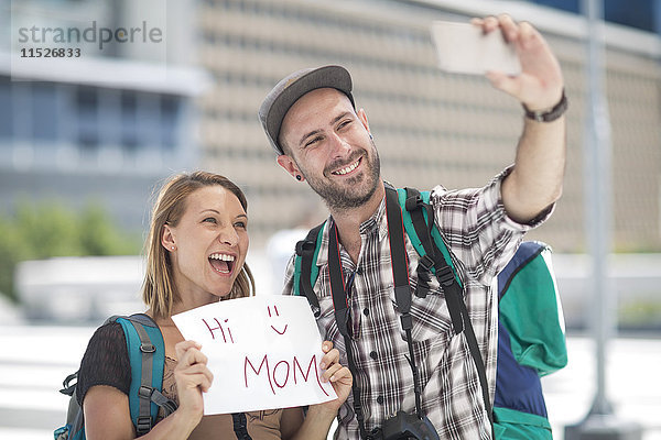 Junges Reisepaar beim Fotografieren mit Schild'Hallo Mama
