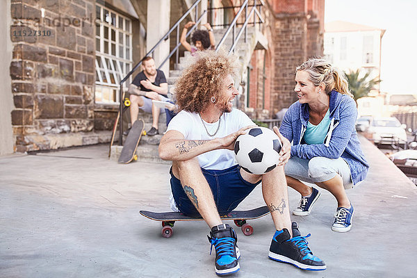 Lächelndes Paar mit Fußball und Skateboard im Gespräch auf dem städtischen Bürgersteig