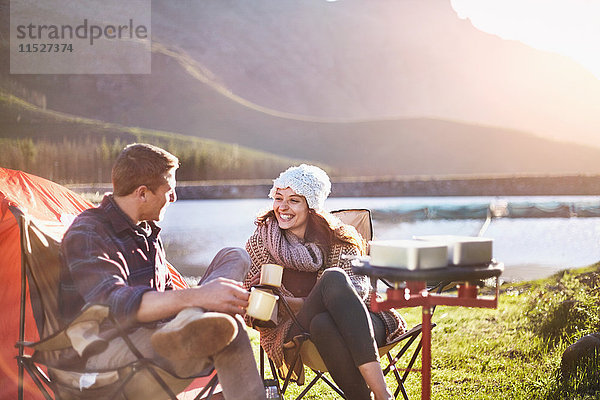 Lächelndes junges Paar trinkt Kaffee auf dem sonnigen Campingplatz am See.