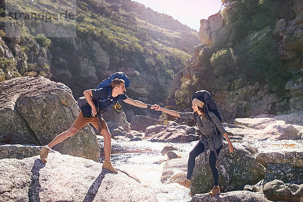 Junger Mann hilft Freundin beim Wandern  überquert sonnige Felsen im Bach