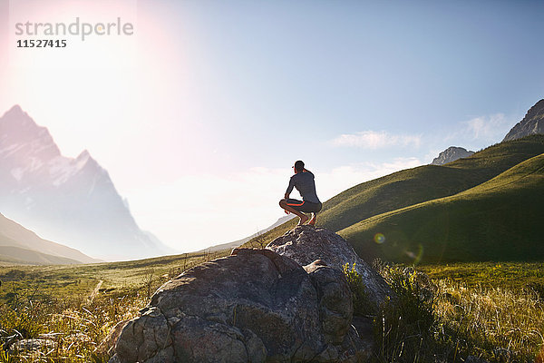 Junger Mann kauernd auf dem Felsen  mit Blick auf sonnige  emote Bergsicht