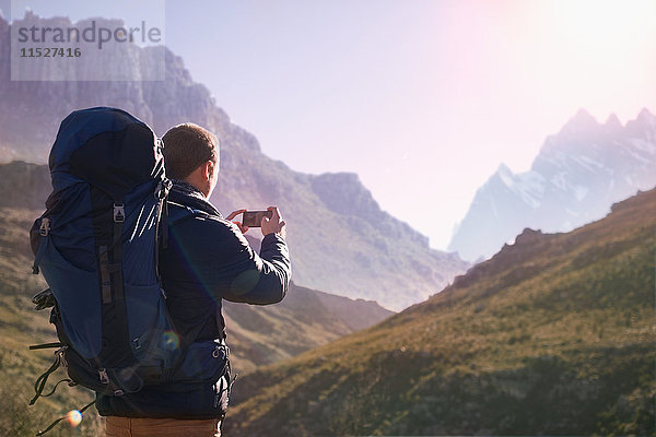 Junger Mann mit Rucksack mit Fotohandy im sonnigen Tal unter den Bergen