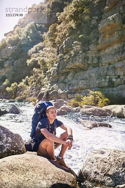 Junger Mann mit Rucksackwanderung  auf Felsen am sonnigen Fluss ruhend