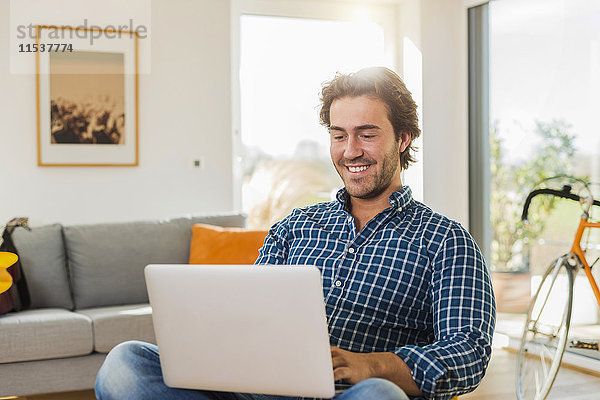Porträt des lächelnden jungen Mannes im Wohnzimmer mit Laptop