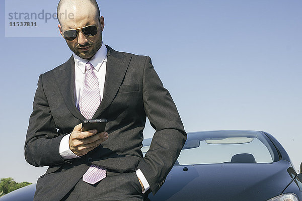 Geschäftsmann mit Sonnenbrille  der sich gegen das Auto lehnt und auf das Handy schaut.