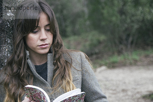 Junge Frau beim Lesen eines Buches  das sich an eine Korkeiche lehnt